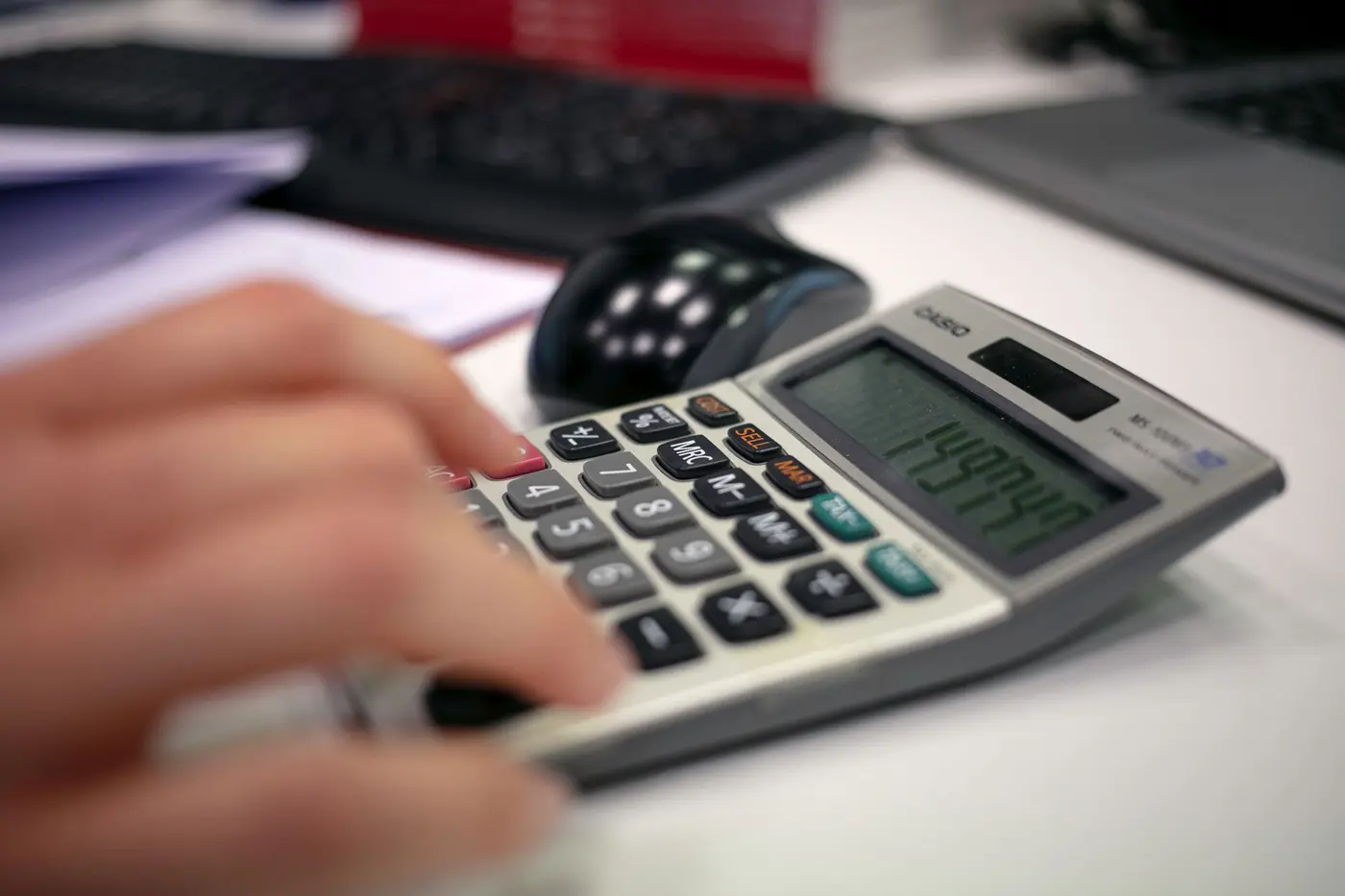 A desk calculator in use beside a keyboard and a stack of paperwork