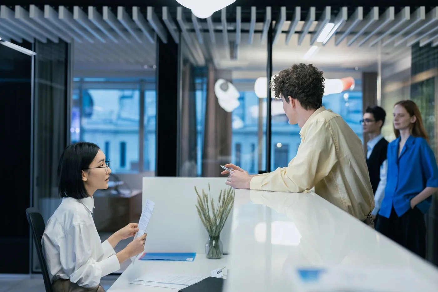 A client leaning on an office front desk talking with the person behind it, two more people waiting nearby