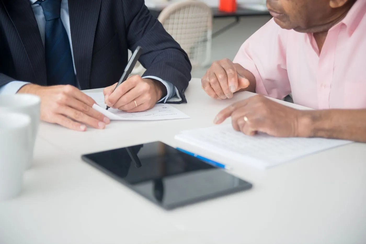Two people at a white table going over a document together, one writing, a tablet and coffee cups between them